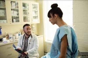 Una mujer transgénero con una bata de hospital hablando con su médico, un hombre transgénero, en una sala de examen.