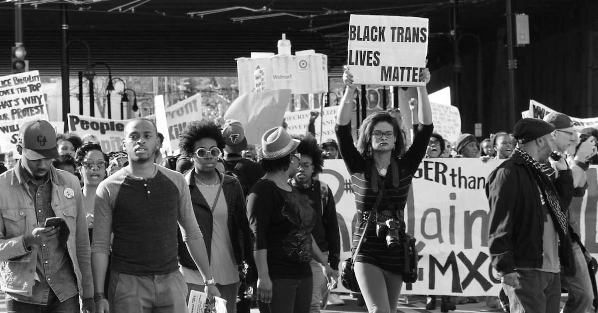 A black-and-white photo of people marching in the street and holding protest signs. The most visible sign says "Black trans lives matter".