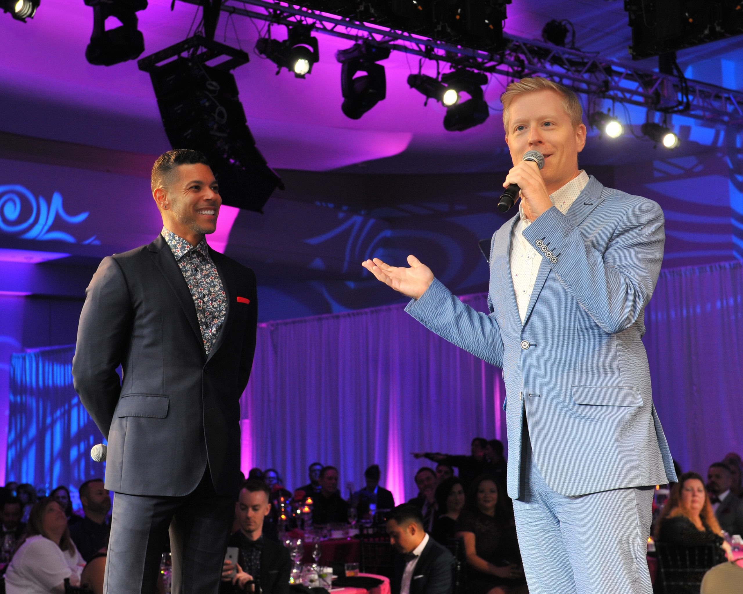 Two people in suits stand on stage, one speaking into a microphone. In the background are tables of gala attendees.