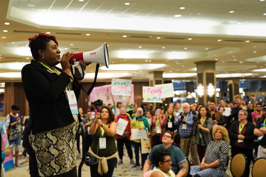 Monica Roberts speaks into a megaphone as a crowd of people holding "Trans Lives Matter" signs stand and sit in the background.