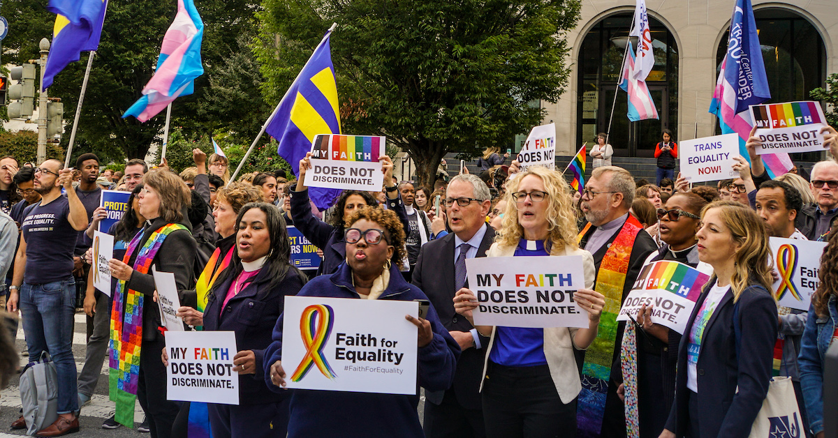 A group of clergy sing in front of a crowd of people rallying while holding signs that say "My Faith Does Not Discriminate," "Fath for Equality," and "Trans Equality Now".