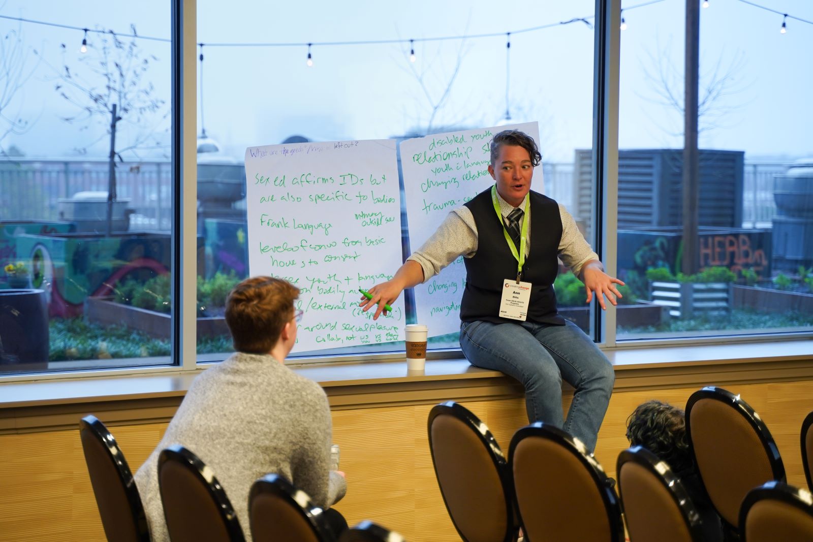 Presenter sits on window ledge next to large sticky paper on window, speaking to participants in chairs