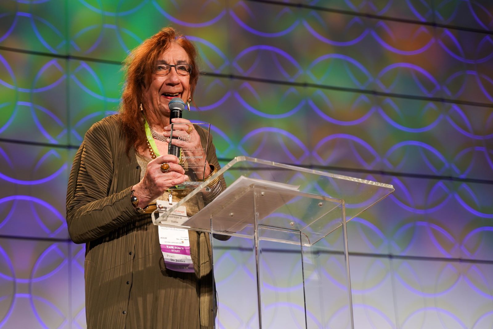 Barbara Satin stands behind a podium holding a microphone, speaking to a large crowd