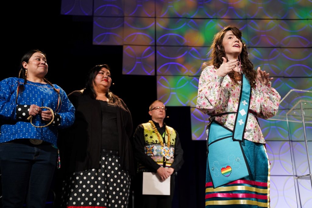 A young Indigenous Leader speaks to a crowd, with three other indigenous people looking on from behind.