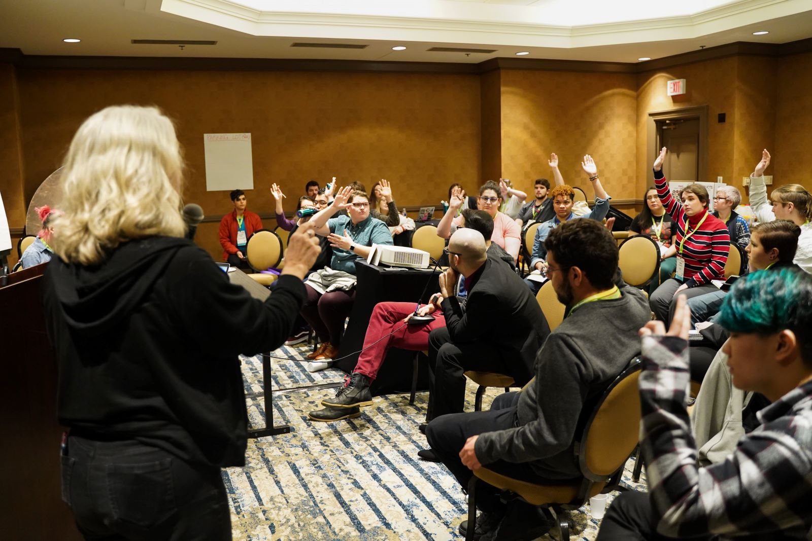 A group of people in a small conference session raise their hands, prompted by the session leader who is raising their hand from the front of the room.