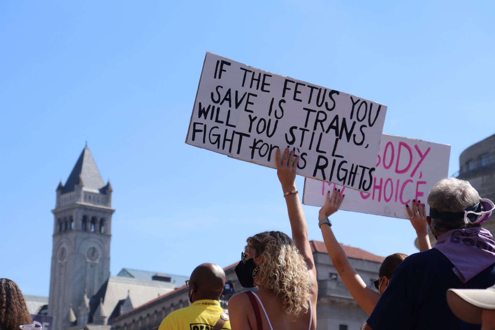 A crowd gathers for a rally for reproductive rights. A woman holds a sign reading "If the fetus you save is trans, will you still fight for its rights?"