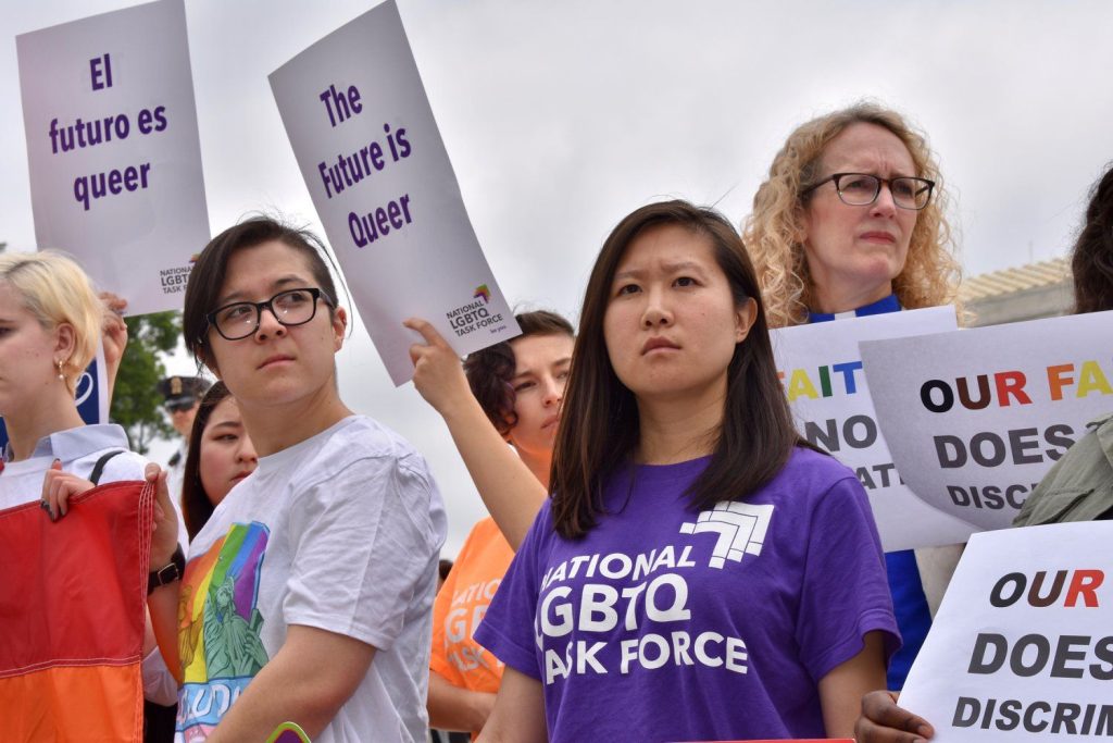 La mujer API con camisa morada se encuentra entre la multitud en el mitin con una mirada determinada en su rostro, vistiendo un morado Task Force Camiseta Nacional LGBTQ. Un cartel al fondo dice "El futuro es extraño".