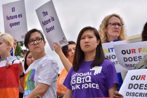 API woman in purple shirt stands in crowd at rally with determined look on her face, wearing a purple National LGBTQ Task Force shirt. A sign in the background reads "The future is queer."