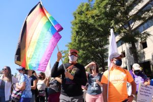 A crowd of people holding protest signs. The person in the foreground is waving a large rainbow flag.