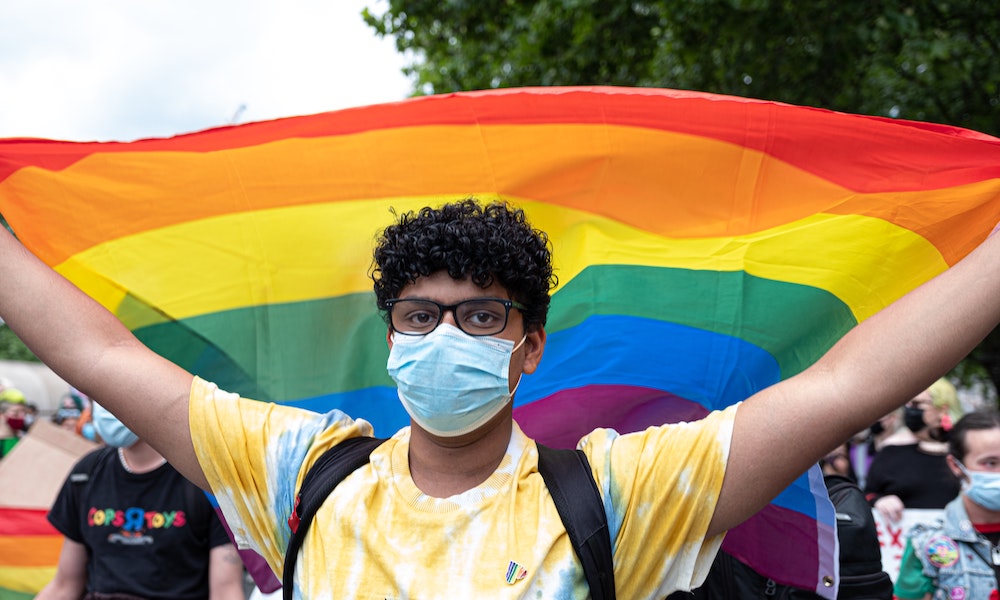 A person looking into the camera, holding a rainbow flag aloft behind them with both hands.