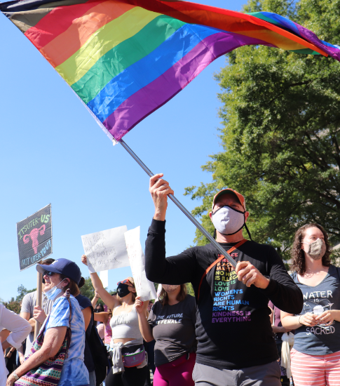 A crowd of people holding protest signs. The person in the foreground is waving a large rainbow flag.