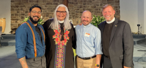 Four people standing in a row, smiling at the camera. The second and fourth people are wearing clerical collars.