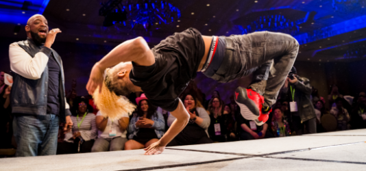 A breakdancer breaking on stage. In the background are the MC and an applauding audience.