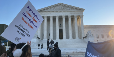 The Supreme Court. In the foreground is a protest sign that says "Repro Rights are Queer Rights. Queer Rights are Repro Rights."