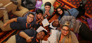 A ground of 7 people lying on the floor in a circle and smiling up at the camera. The person closest to the camera is giving a thumbs-up.