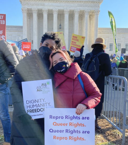 Two people at a rally outside the Supreme Court. They hold signs saying "Dignity. Humanity. Freedom" and "Repro Rights are Queer Rights. Queer Rights are Repro Rights."