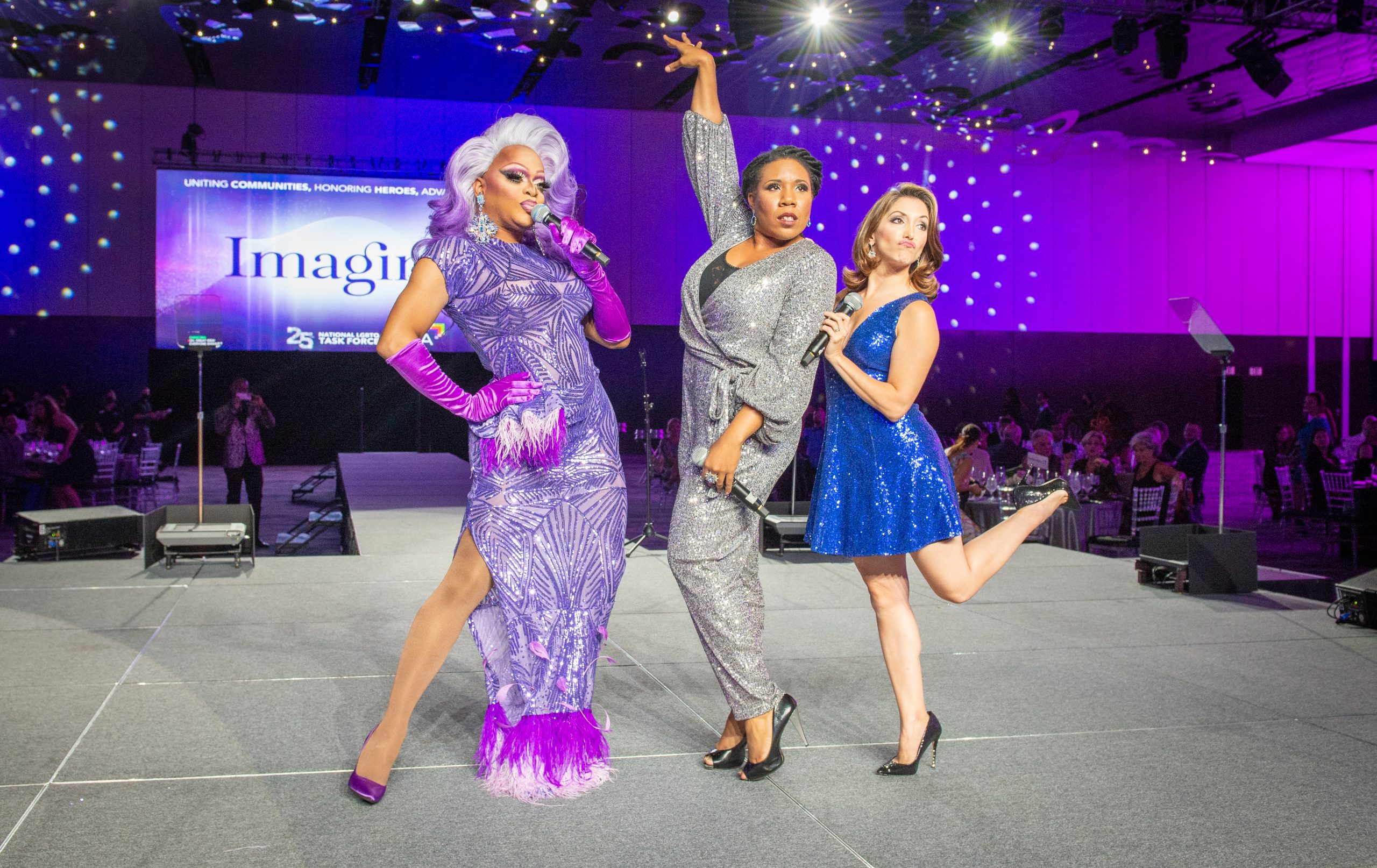 Three people stand on stage striking poses as tables of gala attendees look on.