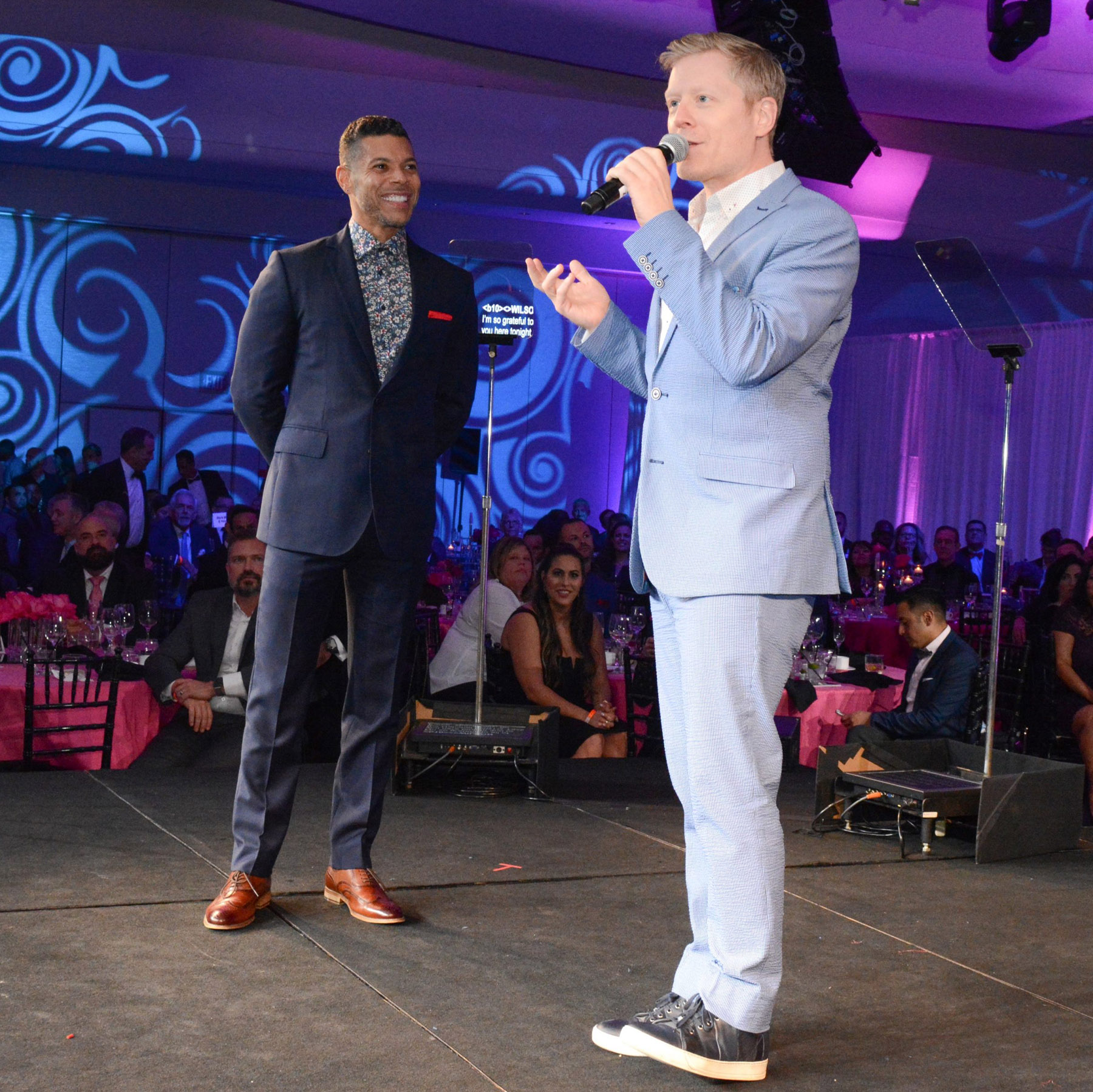 Wilson Cruz and Anthony Rapp stand on stage at the Task Force gala. Rapp is speaking into the microphone.