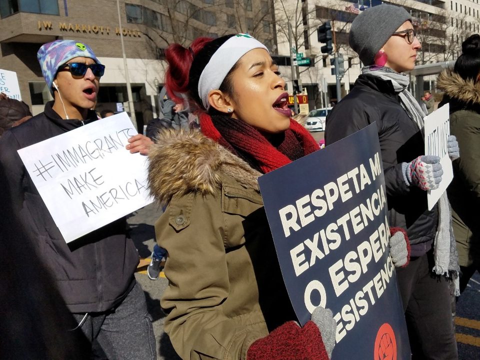 Grupo de defensores de los derechos de los inmigrantes marchan en la calle con abrigos, mitones y sombreros. Llevan carteles que dicen "Los inmigrantes hacen Estados Unidos" y "Respeta mi existencia o Espera Resistencia". Ellos están cantando.