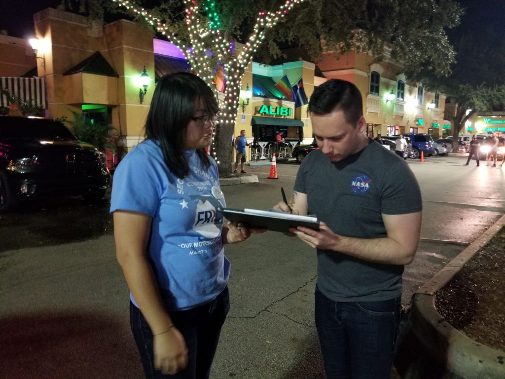 Un encuestador de API que lleva una camisa azul claro que dice "Segundas oportunidades" le entrega un portapapeles a una persona blanca con una camisa gris, que está firmando un papel en el portapapeles. Están de pie en una calle bien iluminada por la noche con banderas de arcoíris en una tienda al fondo.