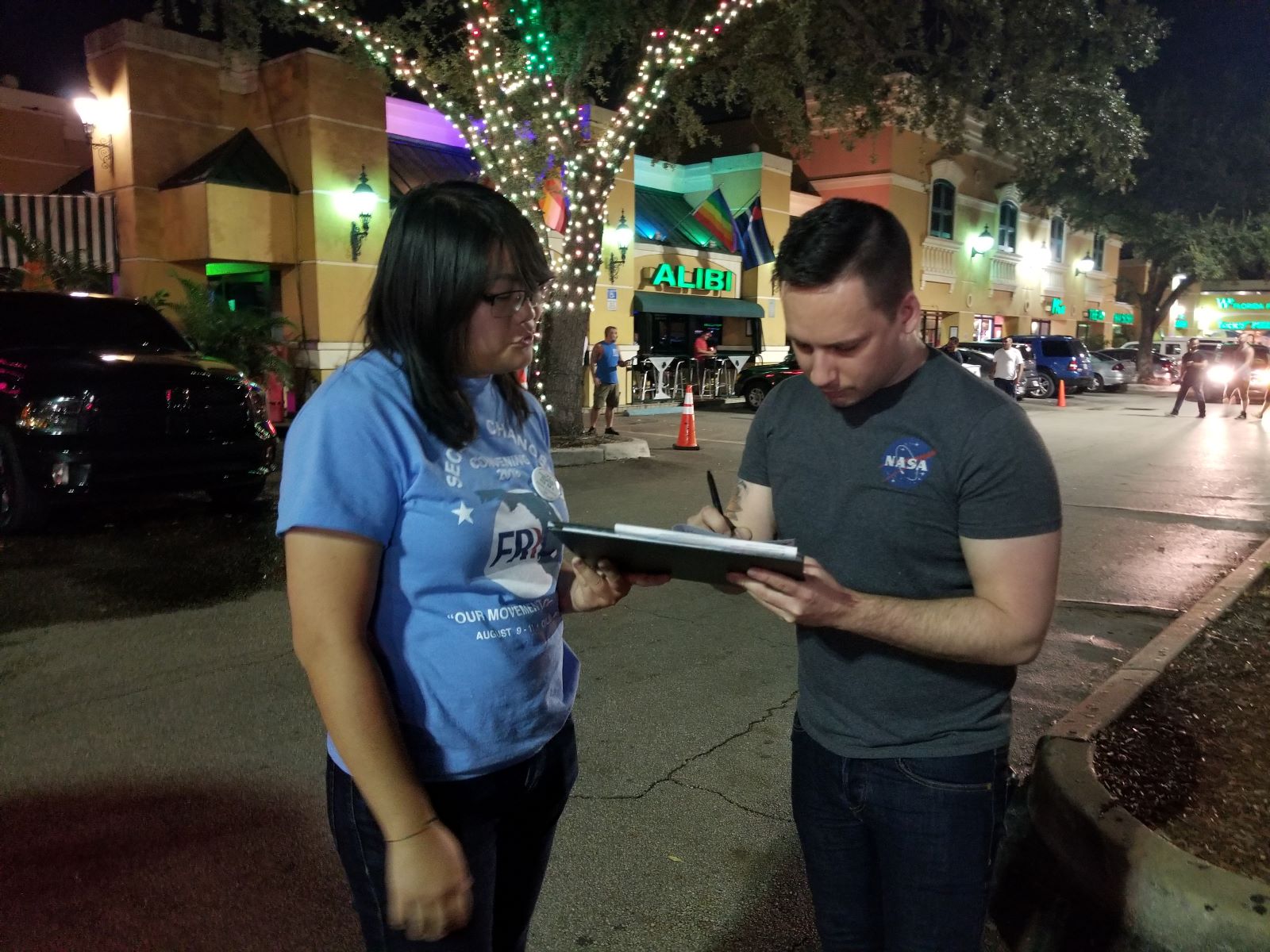 An API canvasser wearing a light blue shirt reading "Second chances" hands a clipboard to a white person in a grey shirt, who is signing a paper on the clipboard. They are standing in a well-lit street at night with rainbow flags on a storefront in the background.