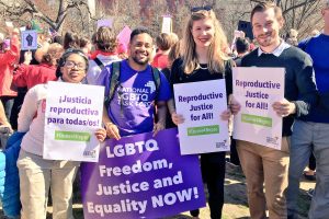 A group of young black and white advocates hold signs at an outdoor gathering. Their signs read "Justicia reproductive para todas/os," "LGBTQ Freedom, Justice and Equality NOW," and "Reproductive Justice for All." The signs include the hashtag #Queer4Repro. All four people are smiling and facing the sun.