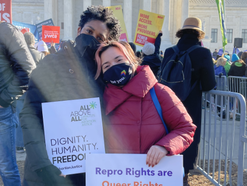 Two people at a rally outside the Supreme Court. They hold signs saying "Dignity. Humanity. Freedom" and "Repro Rights are Queer Rights. Queer Rights are Repro Rights."