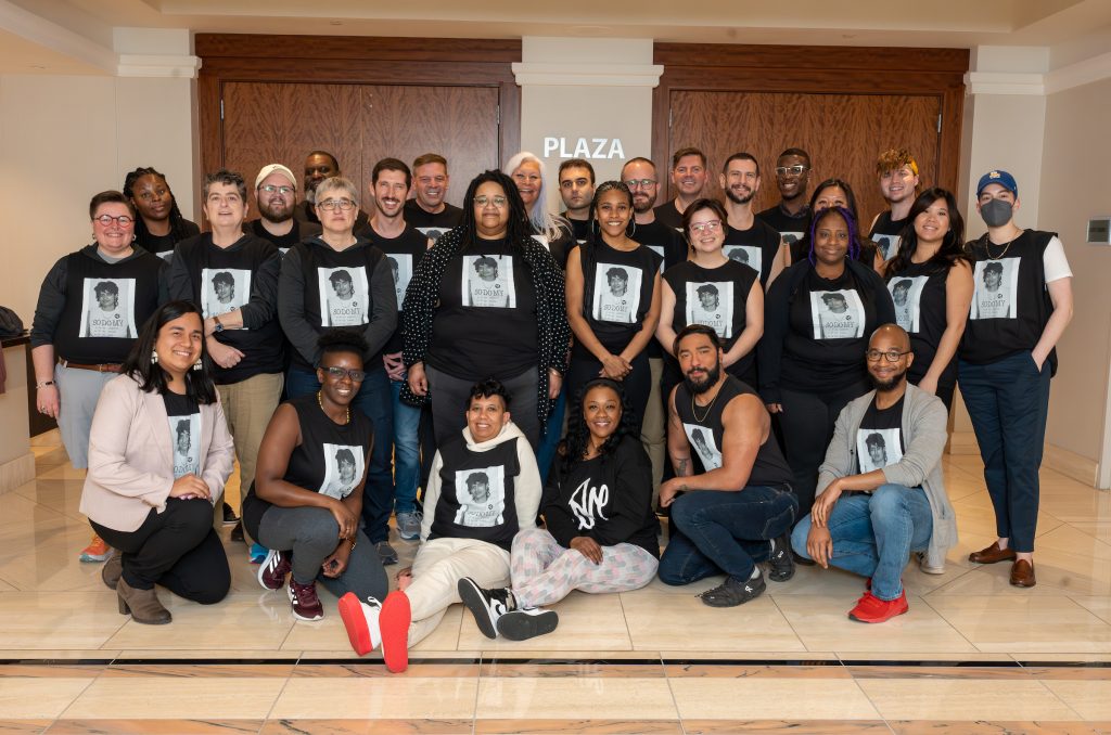 A multi-racial group of 28 LGBTQ+ people gathered in a group posing for a photo. They are all wearing the same shirt featuring a photo of Urvashi Vaid.