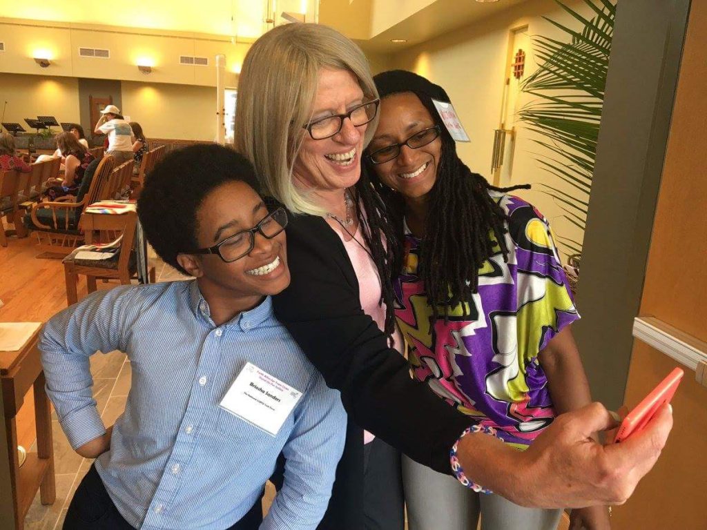 Three queer and transgender individuals stands closely together, smiling. The white woman in the middle holds a camera in her outstretched arm as she takes a selfie of the group. The young black person on the left poses with their hand on their hip, and the young black woman on the right smiles as she leans into the picture.