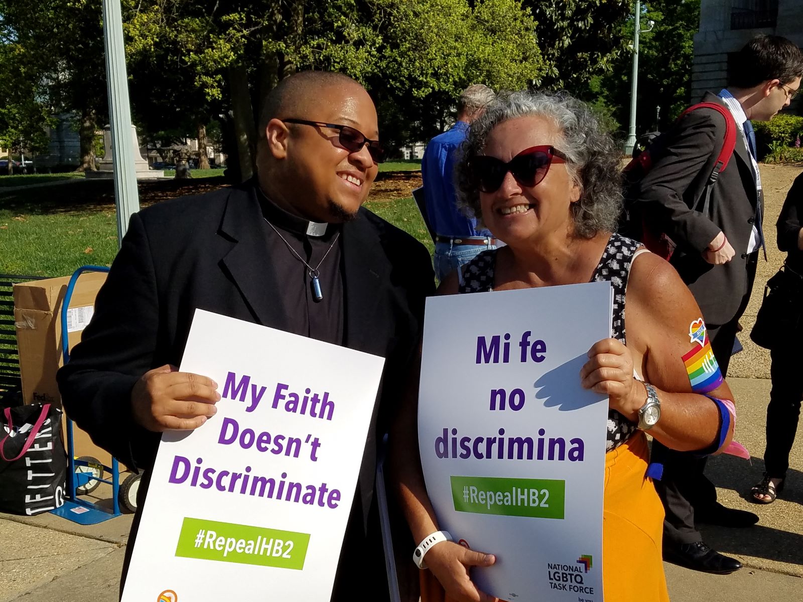 A black transgender man wearing a white clergy collar, black shirt, and black suit jacket holds a sign reading "My Faith Doesn't Discriminate." He is standing outdoors in the sun, wearing sunglasses. He is smiling and looking toward the person next to him, an older white woman who is smiling at the camera. She is wearing a sleeveless shirt and has rainbow stickers on her arm. Her sign reads "Me fe no discrimina." Both signs include the hashtag #RepealHB2