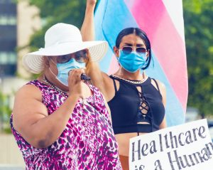 Two trans Latinas at a protest, one woman is holding a microphone and the other holding a trans flag and a sign that says "Healthcare is a Human Right"