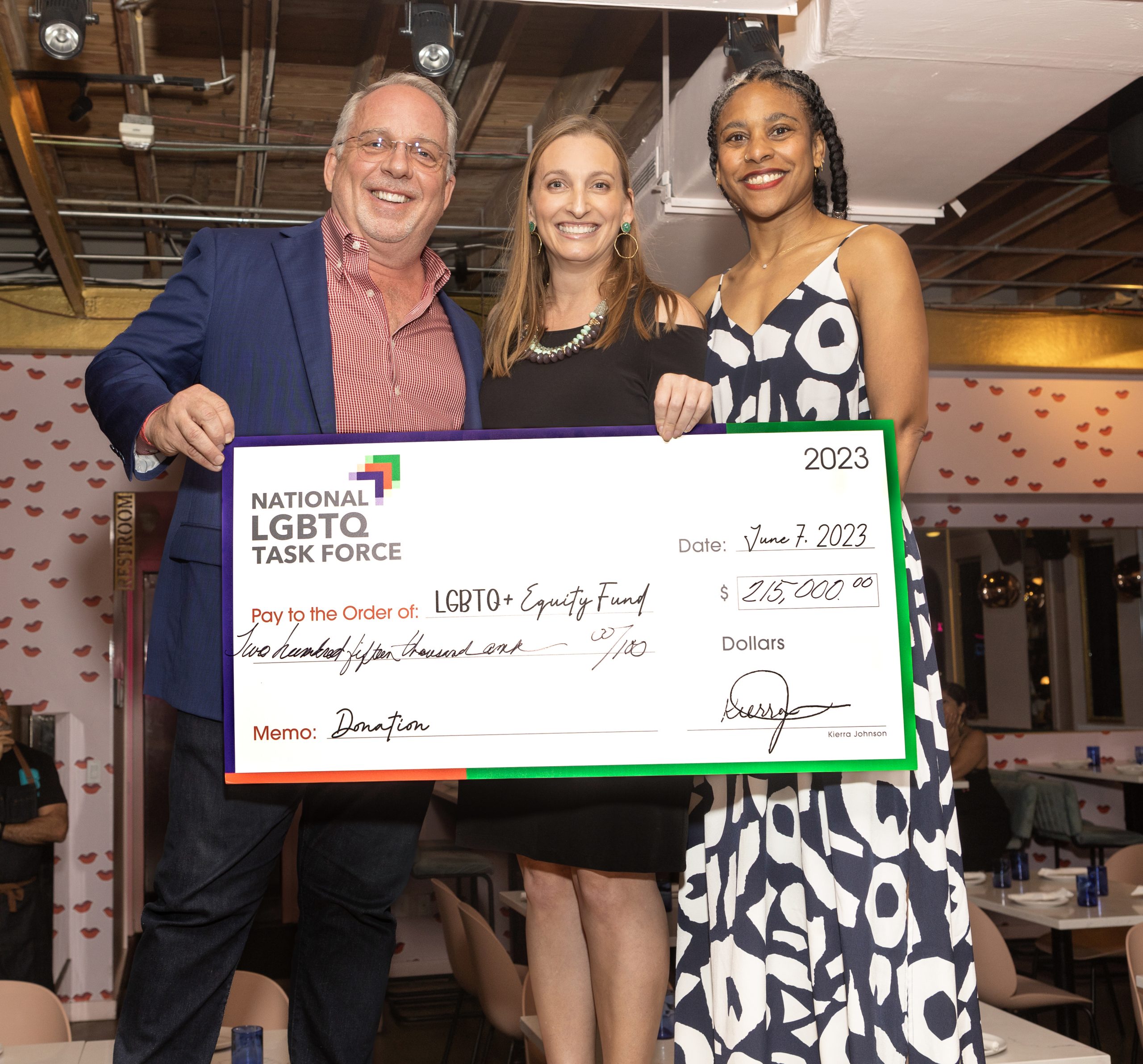 The main focus of the image is on the three people holding a check while participating in an event. They are standing close to each other, and there is a sense of unity and camaraderie. Kierra Johnson, the National LGBTQ Task Force Executive Director, presents a $215,000 check to two LGBTQ+ Equity Fund partners. They are all holding up the check, indicating it is a donation. The check is made out to the LGBTQ+ Equity Fund from the National LGBTQ Task Force. The individuals are all posing and smiling for the camera. Overall, the image captures a moment of joy, unity, and support at the check presentation event. The participants proudly pose with a check, representing a donation or contribution, while other individuals and elements add to the positive atmosphere of the scene.