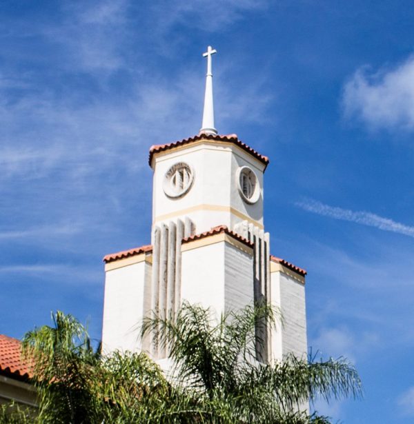 This is a photo of St. John’s on the Lake United Methodist Church in Miami Beach, Florida. The image depicts a scenic view with the church's clock tower, palm trees, and a blue sky. The clock tower is located in the center of the image and stands tall against the backdrop of a clear blue sky. Palm trees can be seen surrounding the clock tower, adding to the tropical feel of the scene.