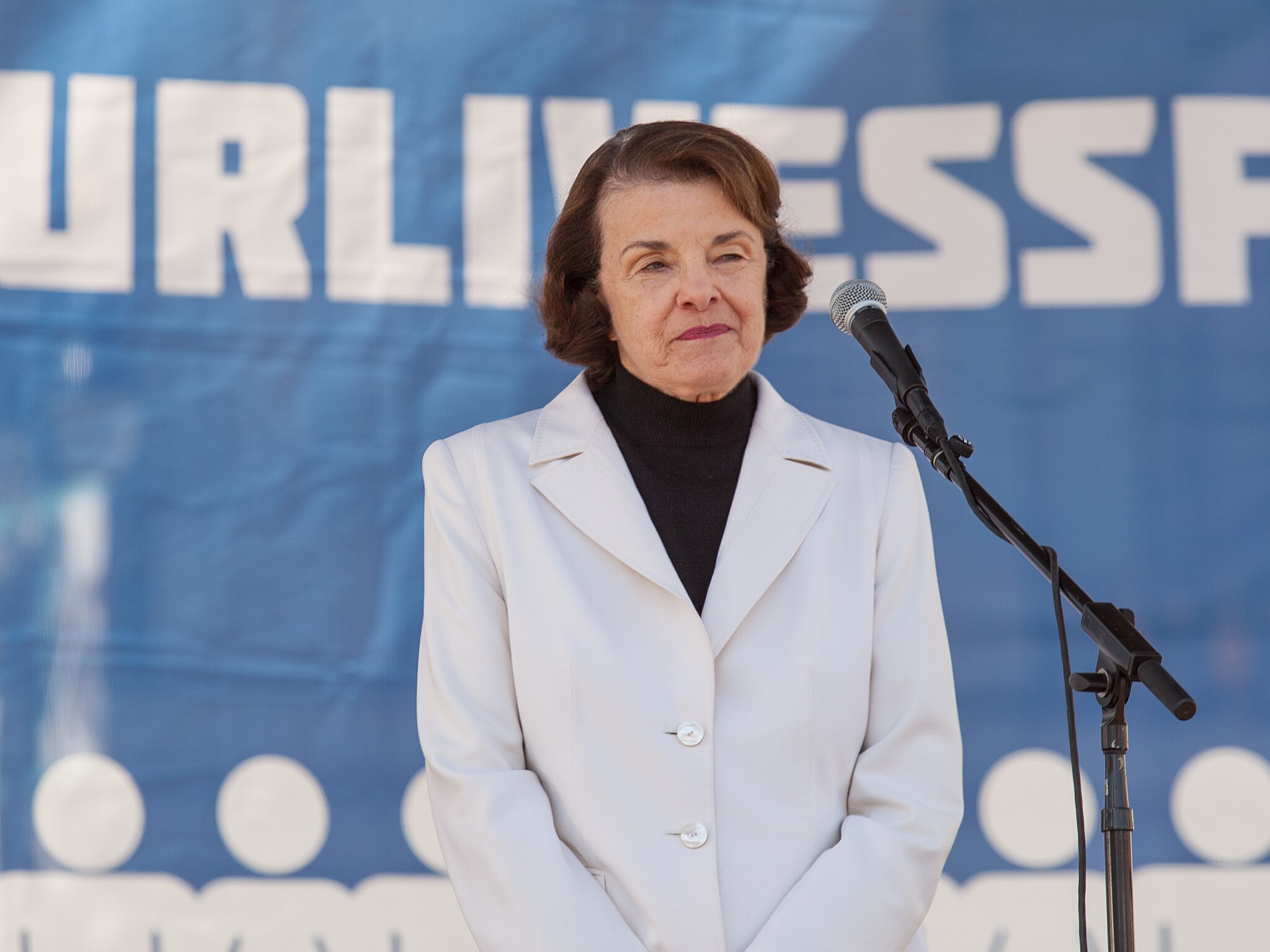 The image features U.S. Senator Dianne Feinstein, an older white woman wearing a white jacket, standing in front of a microphone at the March 2018 San Francisco March for Our Lives rally. She is positioned prominently in the center of the image and is the scene's focal point. Sen. Feinstein is delivering a speech or presentation, as indicated by her proximity to the microphone and engaged expression. Sen. Feinstein is wearing a white suit coat with a black turtle neck under it. The microphone is positioned against a blue background, which adds a contrasting color to the image's predominantly white and neutral tones.