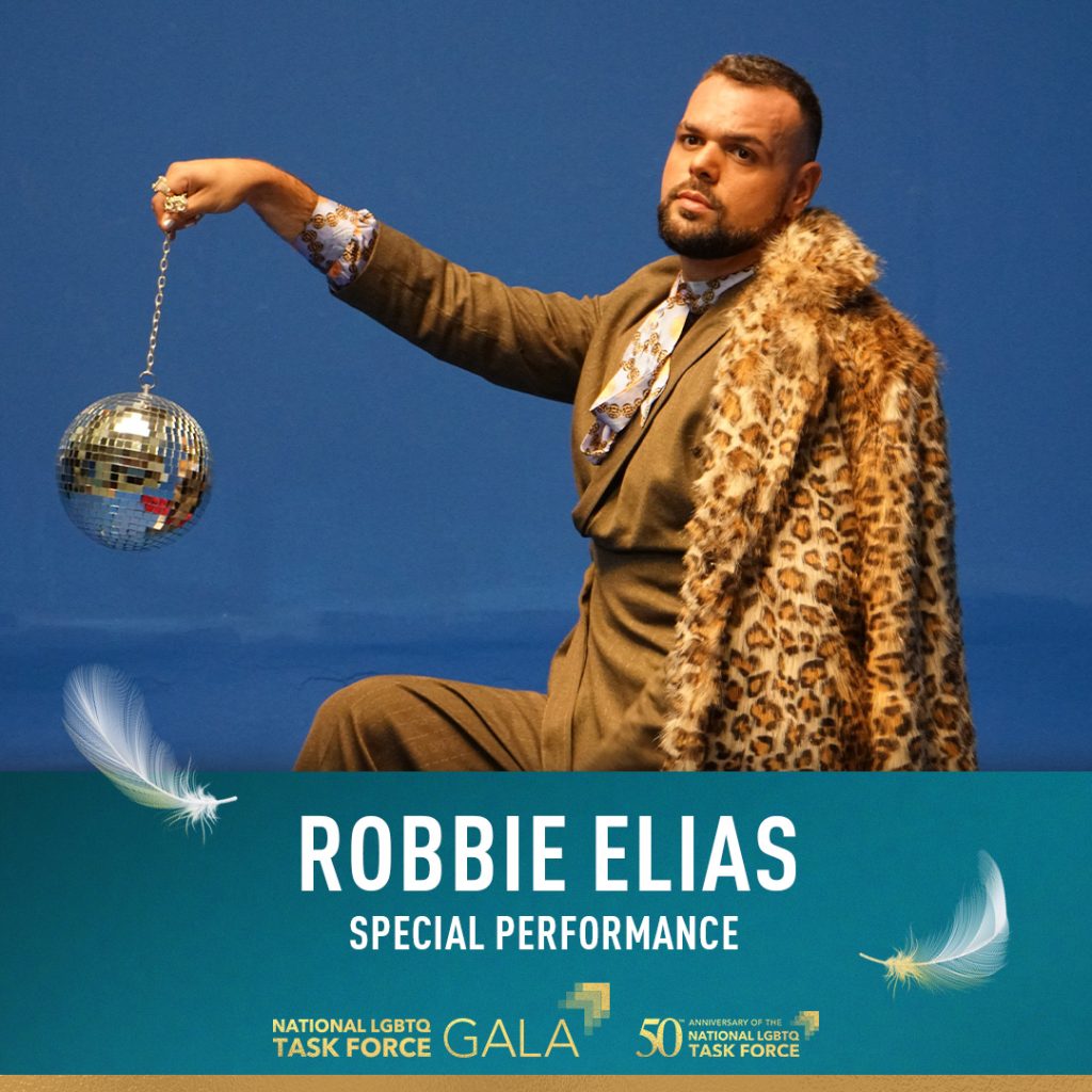 This image features 2023 National LGBTQ Task Force Special Performer Robbie Elias. Robbie Elias is shown wearing a brown suit with an ascot and a leopard print coat hanging off his shoulder. He is standing in the center of the image, exuding confidence. He seems ready to entertain the audience. In the background, there is a disco ball hanging from a string against a blue background. The disco ball reflects light and adds a festive atmosphere to the scene. At the bottom of the image, white text says "Robbie Elias Special Performance" displayed against a blue background. It is positioned towards the center of the image. Towards the bottom right corner of the image, there is text presented in a gradient style, consisting of the words "National LGBTQ Task Force Gala" and "50th Anniversary of the National LGBTQ Task Force." These texts are displayed on a colorful background, with the colors changing from left to right.