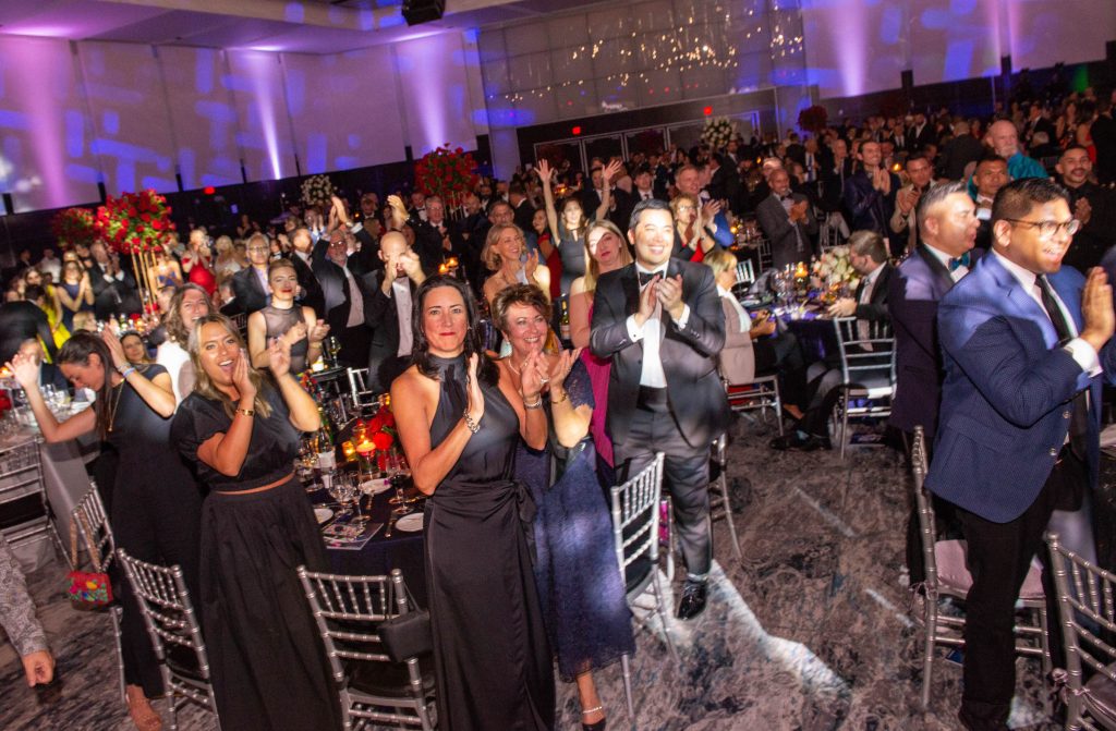 The energetic atmosphere of the 2022 National LGBTQ Task Force Gala is captured in this photograph. A diverse group of individuals has gathered to celebrate and applaud an event. The crowd's enthusiastic clapping fills the room, their applause resonating with a sense of excitement and enjoyment. The attendees are dressed in a variety of attire, with one gentleman standing out in a suit and another in a tuxedo. Among the attendees is a woman wearing a striking black dress, adding an elegant touch to the occasion. The vibrant and dynamic scene is filled with laughter and camaraderie as the guests revel in the festivities. The composition of the photograph creates a sense of unity and togetherness, with the participants forming the centerpiece of the event.