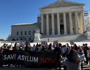 A group of demonstrators hold a large banner that reads "Save Asylum Now" in front of the Supreme Court.