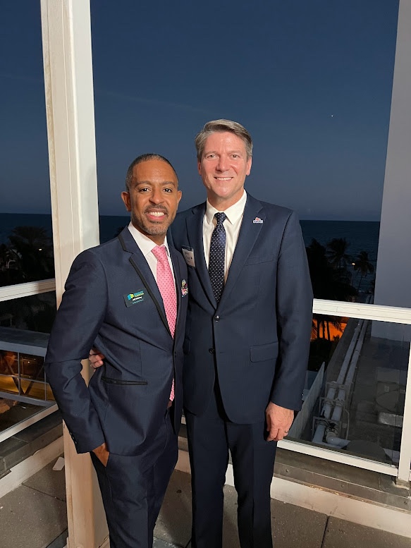 Two men wearing suits (Jeff and Todd Delmay) are standing closely next to each other, presumably on a balcony with a view of the sea at night. The man on the left, Jeff Delamy, is Black and is dressed in a navy blue suit with a pink tie, while the man on the right, Todd Delmay, is White, wearing a navy blue suit, a white shirt, and a blue tie. Behind them, there's a dark blue night sky. This setting suggests that they are at an evening event.