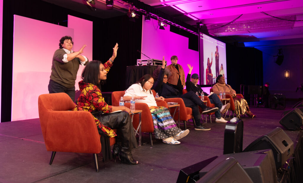 Main stage panel at the Creating Change Conference with speakers raising a hand and engaging with the audience as Kierra Johnson speaks.