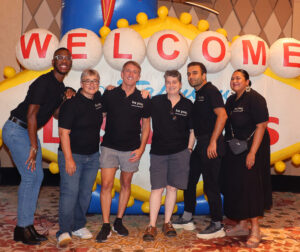 A group of Task Force staff wearing black polo shirts with the slogan "Be You" are standing in front of a blow up figure of the famous landmark sign that reads "Welcome to Fabulous Las Vegas".