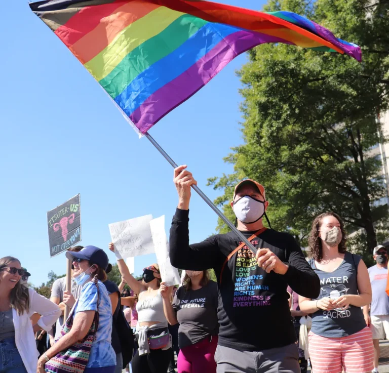 A crowd of people holding protest signs. The person in the foreground is waving a large rainbow flag.